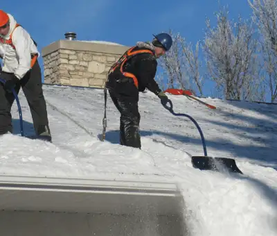 Professional technicians performing roof snow removal with safety harnesses and shovels in Madison, WI